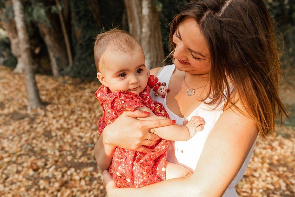 Femme souriante tenant un bébé en robe rouge dans un environnement naturel.